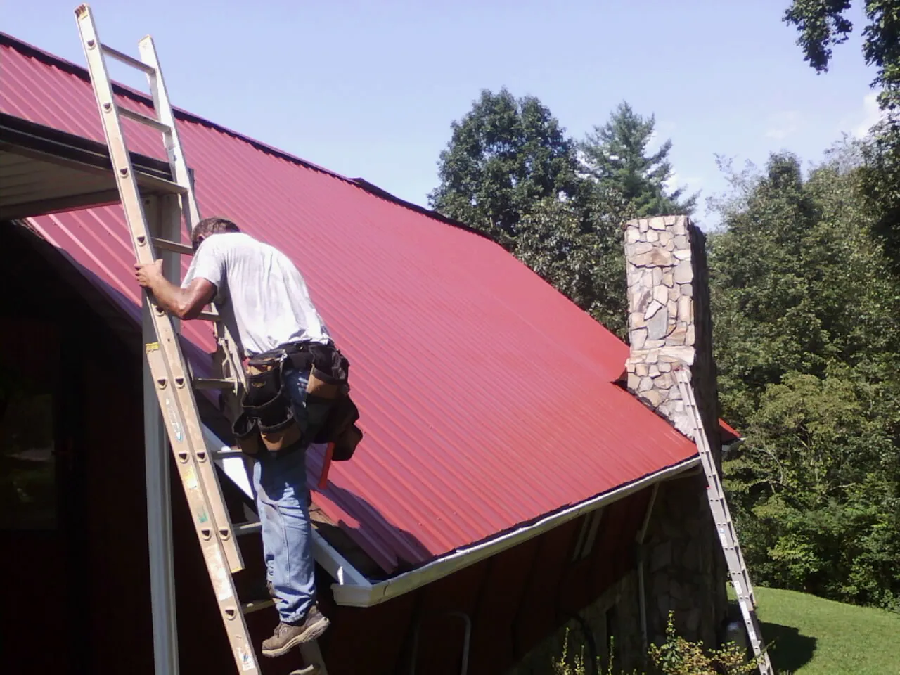 Roofer on a ladder beside a red metal roof and stone chimney — Soffit & Fascia Repair by Roofing Brothers in Monessen